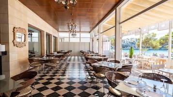 A restaurant with checkered flooring, wicker chairs, and tables set for dining. Large windows offer a view of the sea and a balcony area.