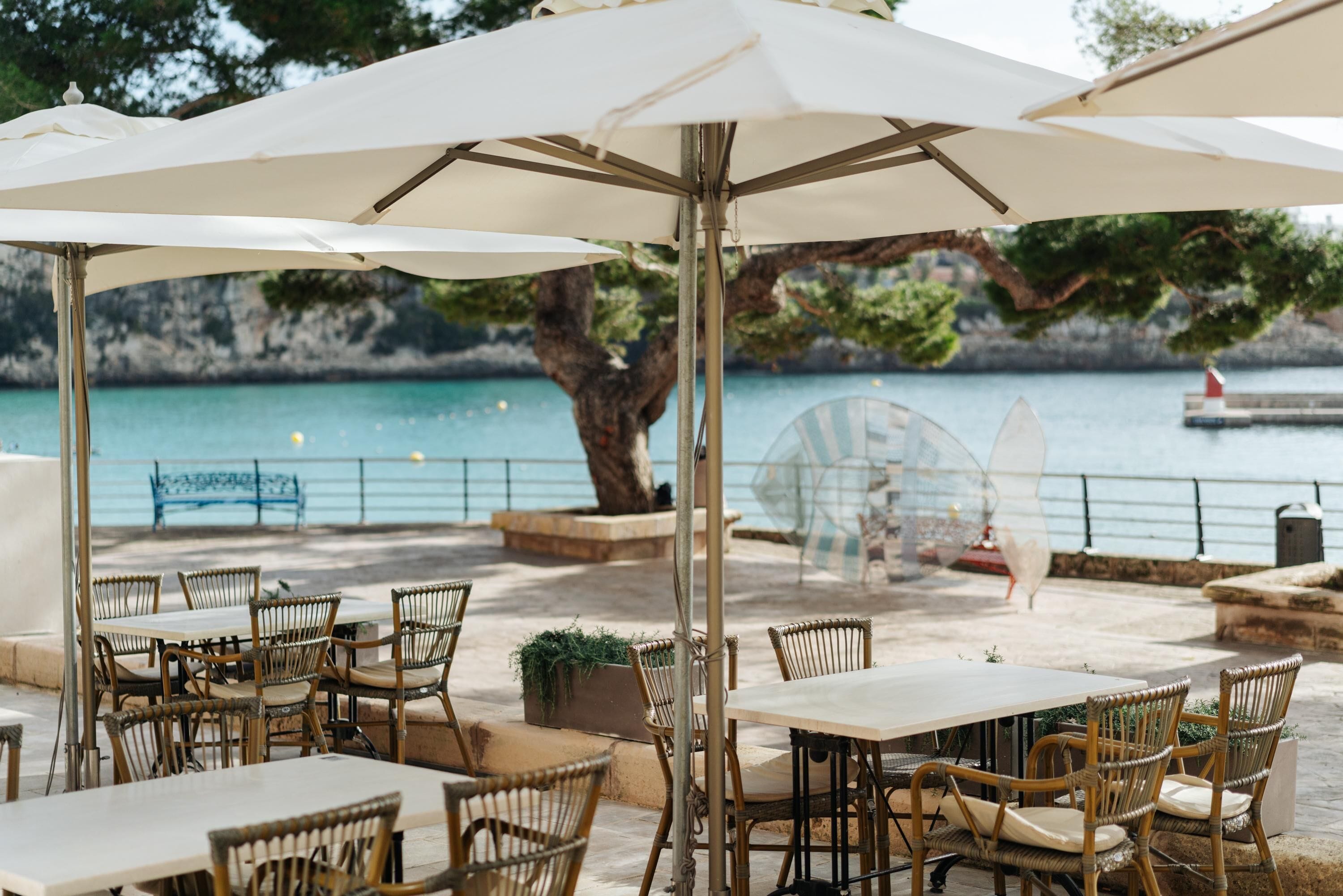 An outdoor seating area with tables and wicker chairs, overlooking a body of water and a rocky coastline.