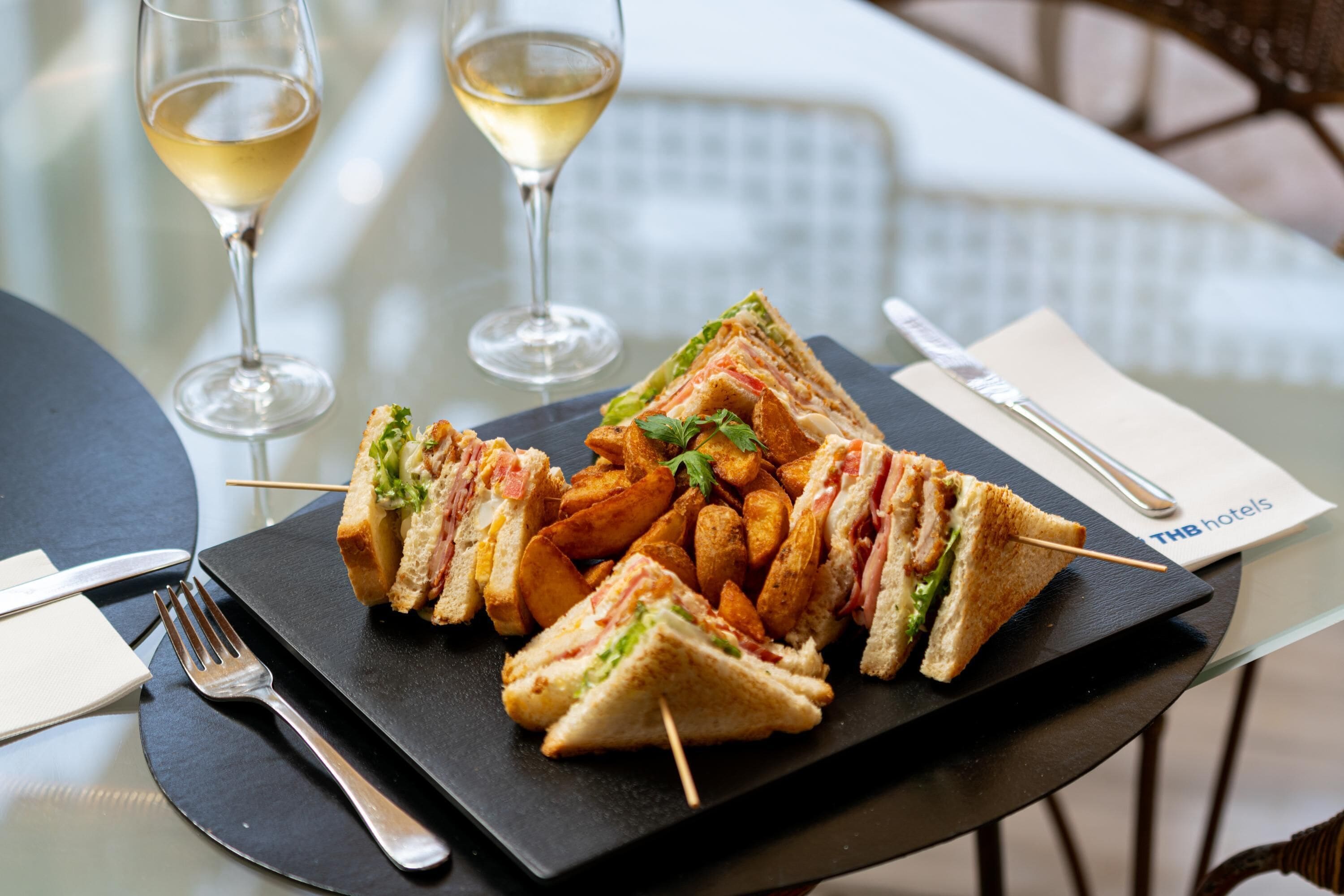 Een tafel met een bord belegd brood, friet, twee wijnglazen, bestek en een servet van THB hotels.