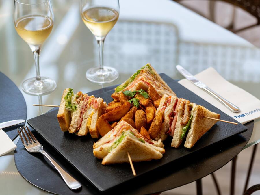 A table with a plate of sandwiches and fries, two wine glasses, cutlery, and a THB hotels napkin.