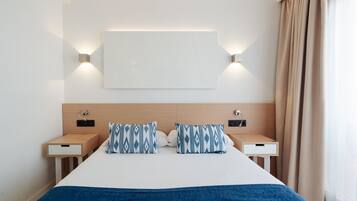 Una habitación de hotel moderna con una cama grande, dos mesitas de noche, una lámpara de pared y un ventanal con cortinas.