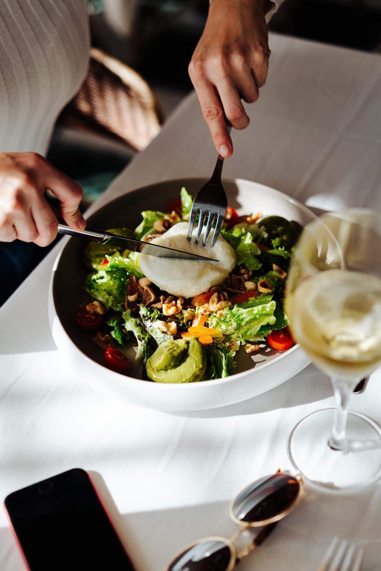 En person sitter vid ett bord och äter en sallad med hjälp av en gaffel, och har ett glas vitt vin framför sig.