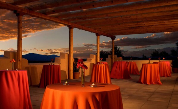 Un espacio para comer al aire libre con mesas redondas cubiertas de manteles rojos, con el cielo del atardecer de fondo.