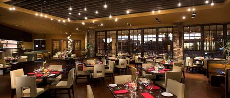 A spacious restaurant with multiple dining tables and chairs, large windows, and a modern ceiling design.