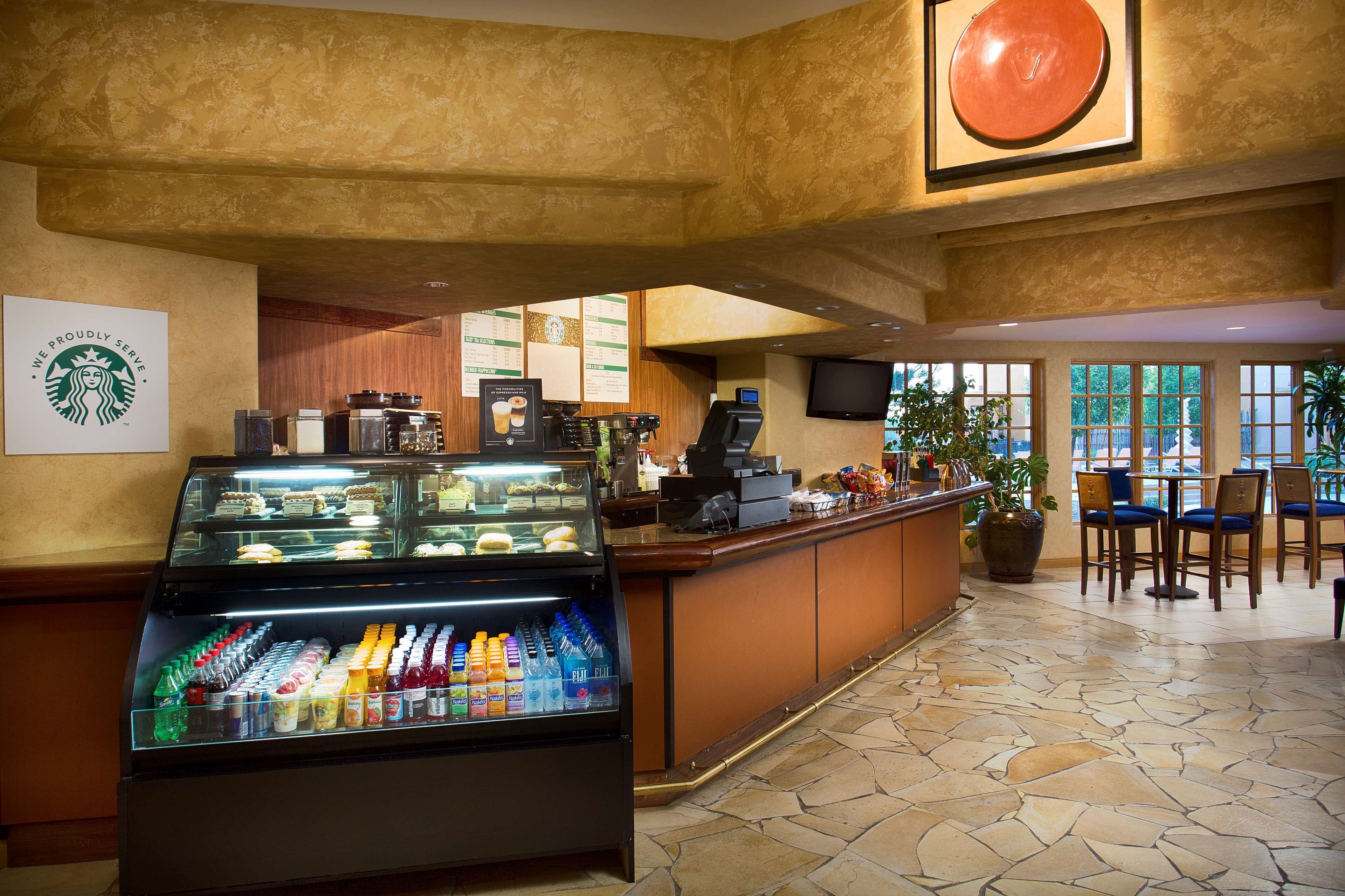 Una cafetería Starbucks con una vitrina de bebidas y pasteles, un mostrador con una caja registradora y una zona de estar con sillas y mesas.