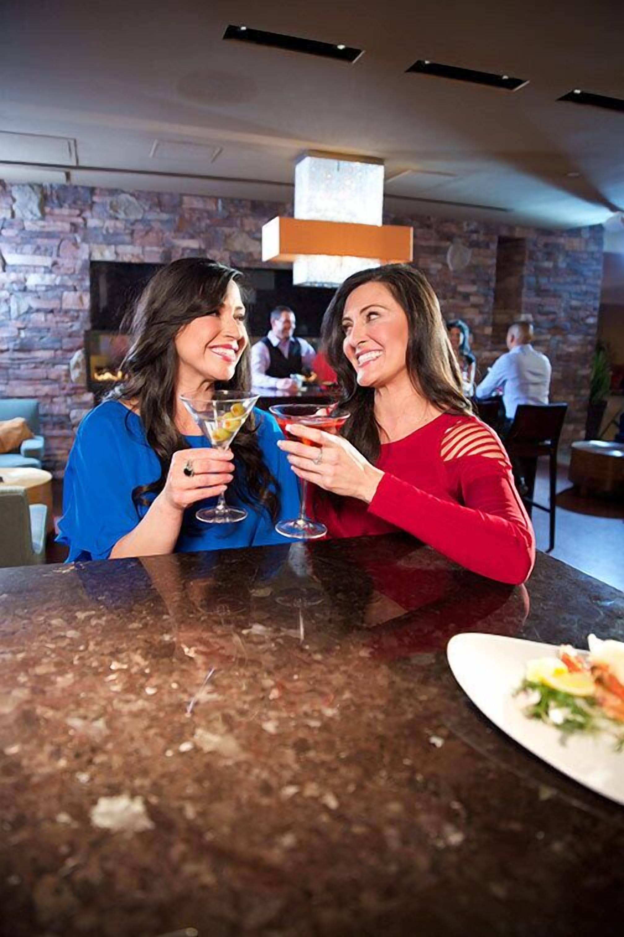 Dos personas brindan con bebidas en un bar con pared de piedra y una lámpara moderna.