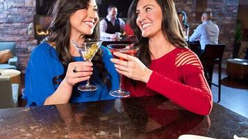 Dos personas brindan con bebidas en un bar con pared de piedra y una lámpara moderna.