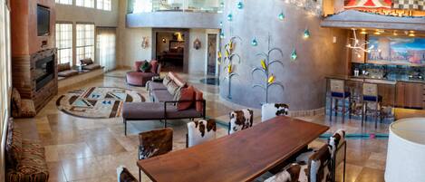 A spacious living area with a large wooden dining table, leather sofas, and a high ceiling with exposed beams.