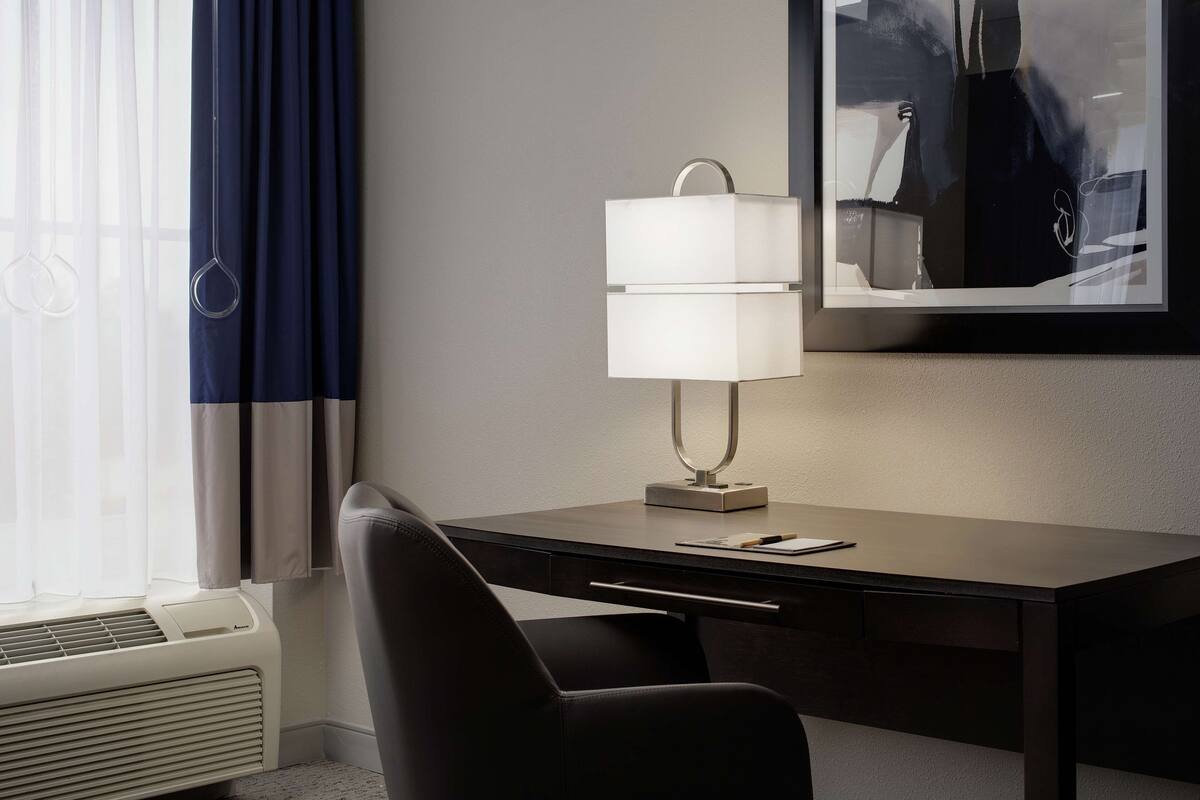 Una habitación de hotel con escritorio, lámpara, silla y ventana con cortinas.