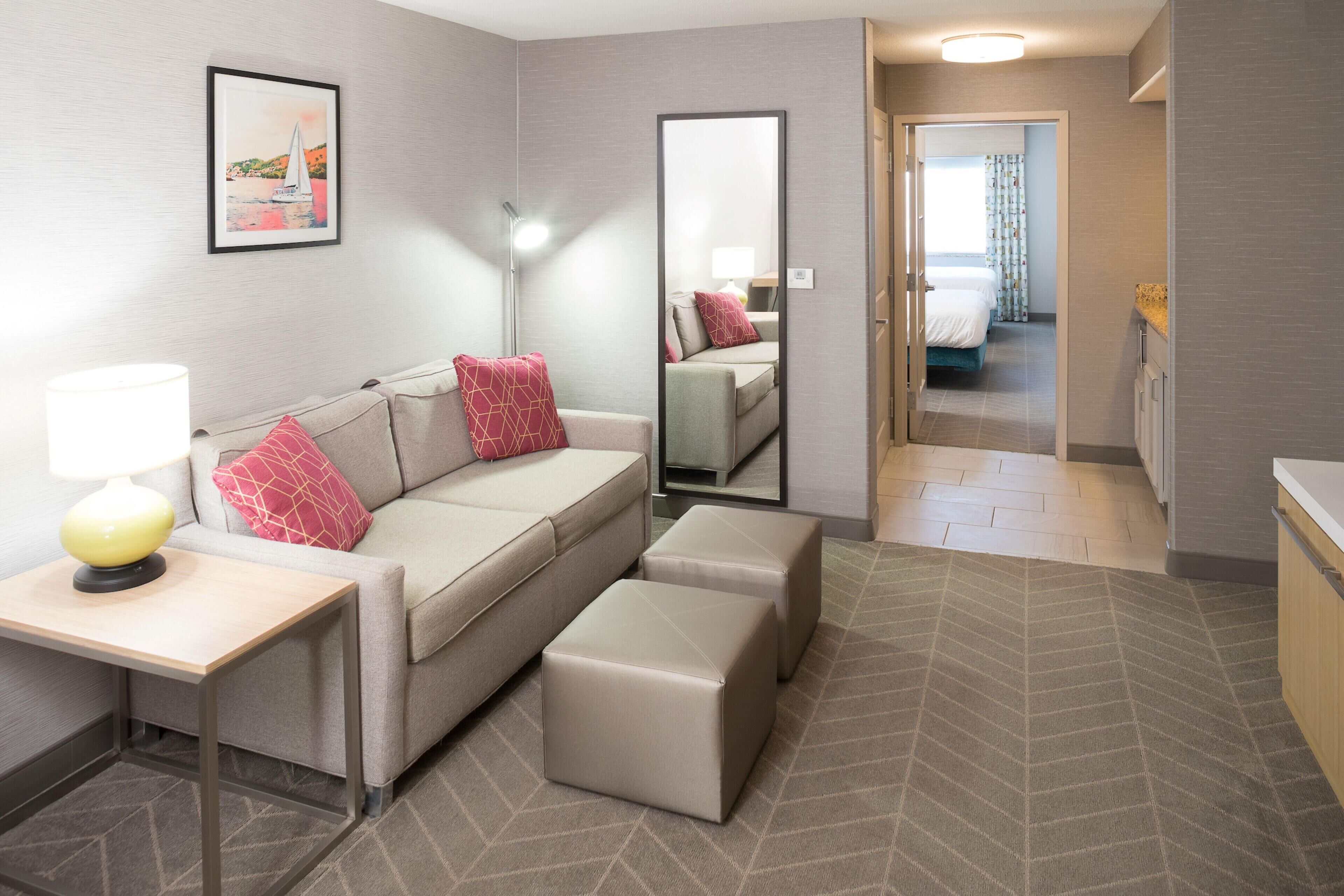 Habitación de hotel con sofá, reposapiés, mesita de noche, lámpara, espejo y vistas al dormitorio contiguo.