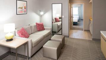 Habitación de hotel con sofá, reposapiés, mesita de noche, lámpara, espejo y vistas al dormitorio contiguo.