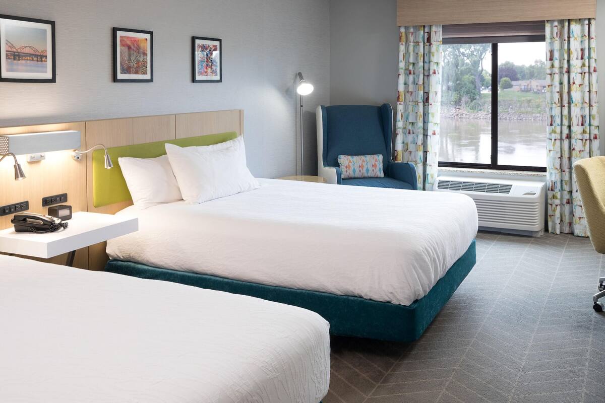 Ein Hotelzimmer mit zwei Betten, einem Schreibtisch mit Telefon, einem Sessel, einer Lampe und einem Fenster mit Vorhängen.
