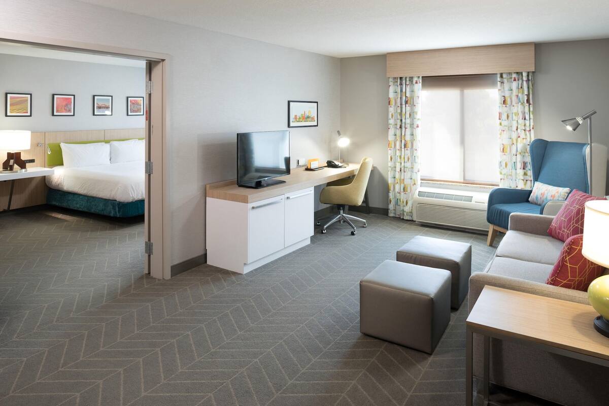 Ein Hotelzimmer mit Bett, Schreibtisch, Stuhl, Hocker und Fernseher.