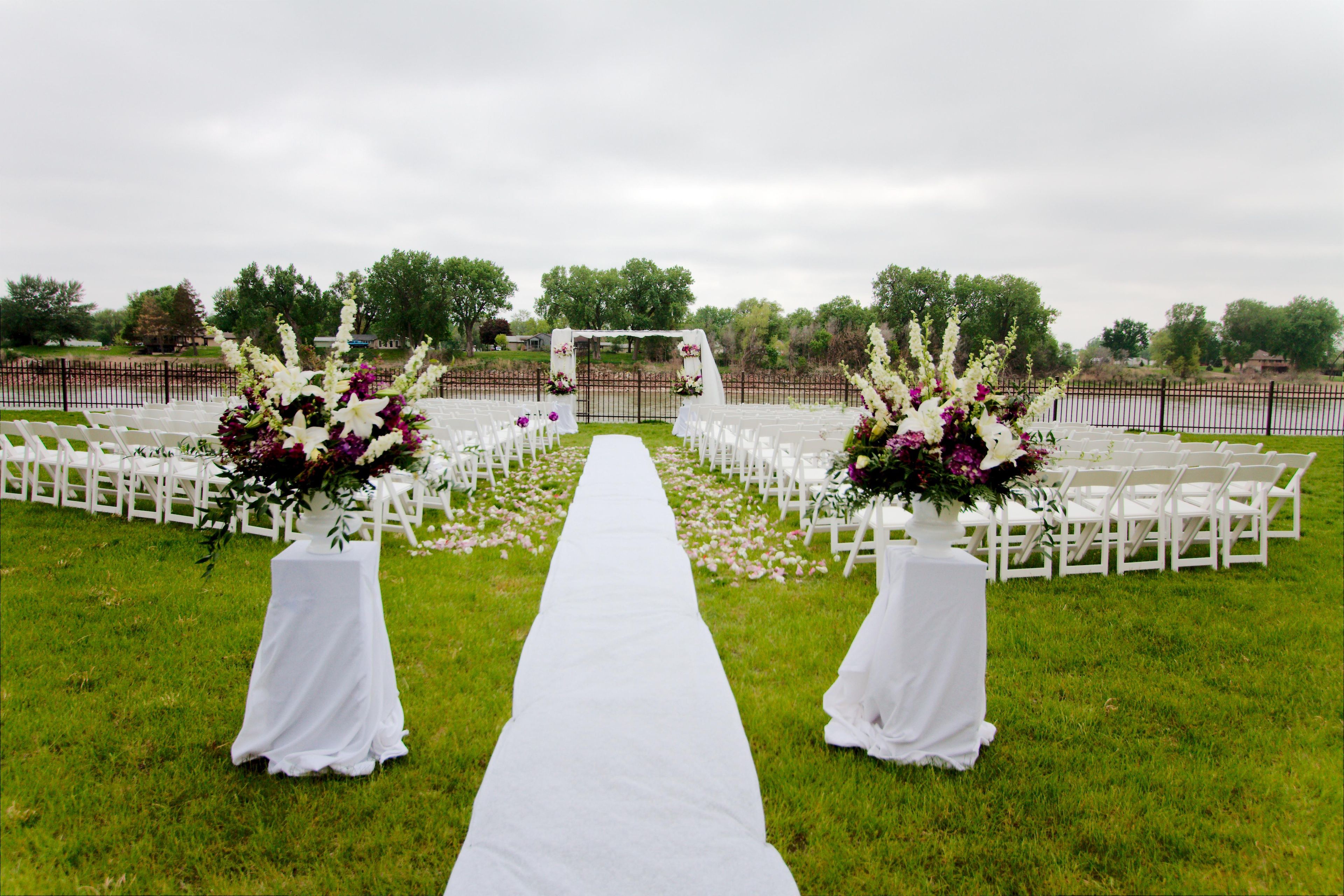 An outdoor wedding aisle with white chairs on either side, a white aisle runner, and floral arrangements at the aisle ends.