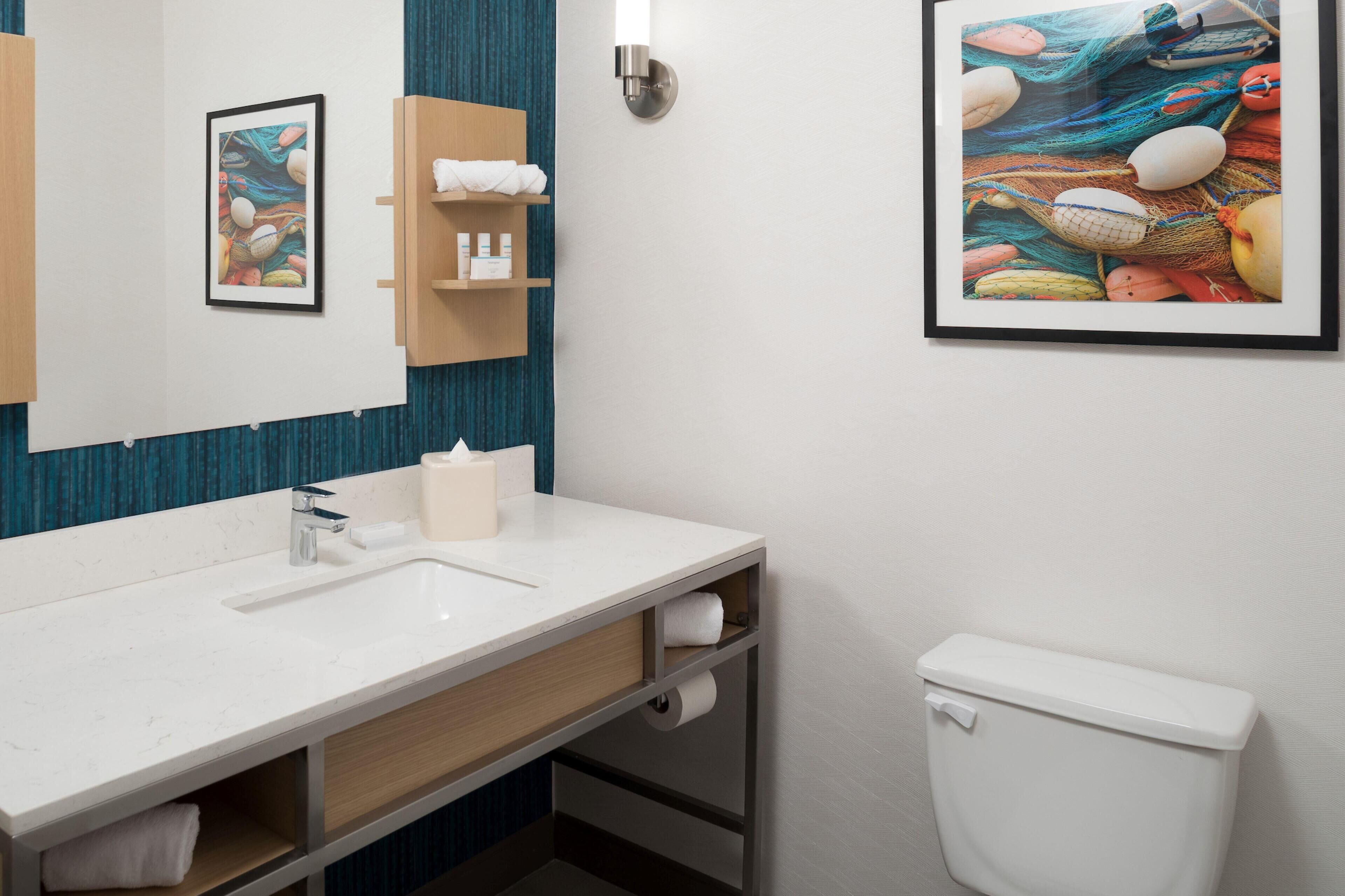 A modern bathroom with a white sink, a toilet, a towel rack, a framed picture, and a shelf with toiletries.