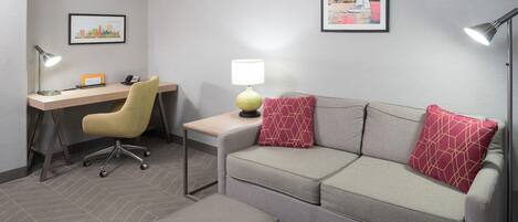 Quarto de hotel com escrivaninha, cadeira, sofá e banquinho. Há duas luminárias, um quadro emoldurado na parede e uma mesinha com luminária.