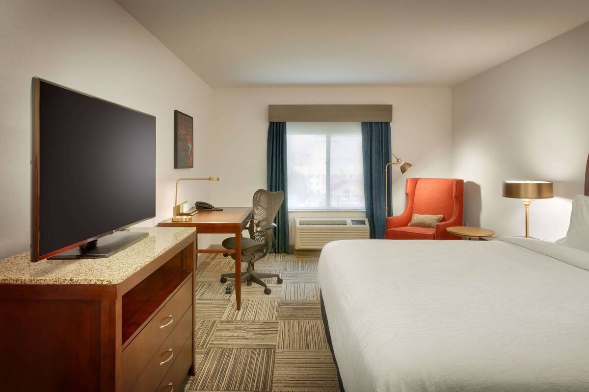 Habitación de hotel con cama, escritorio, silla, televisor y ventana con cortinas.