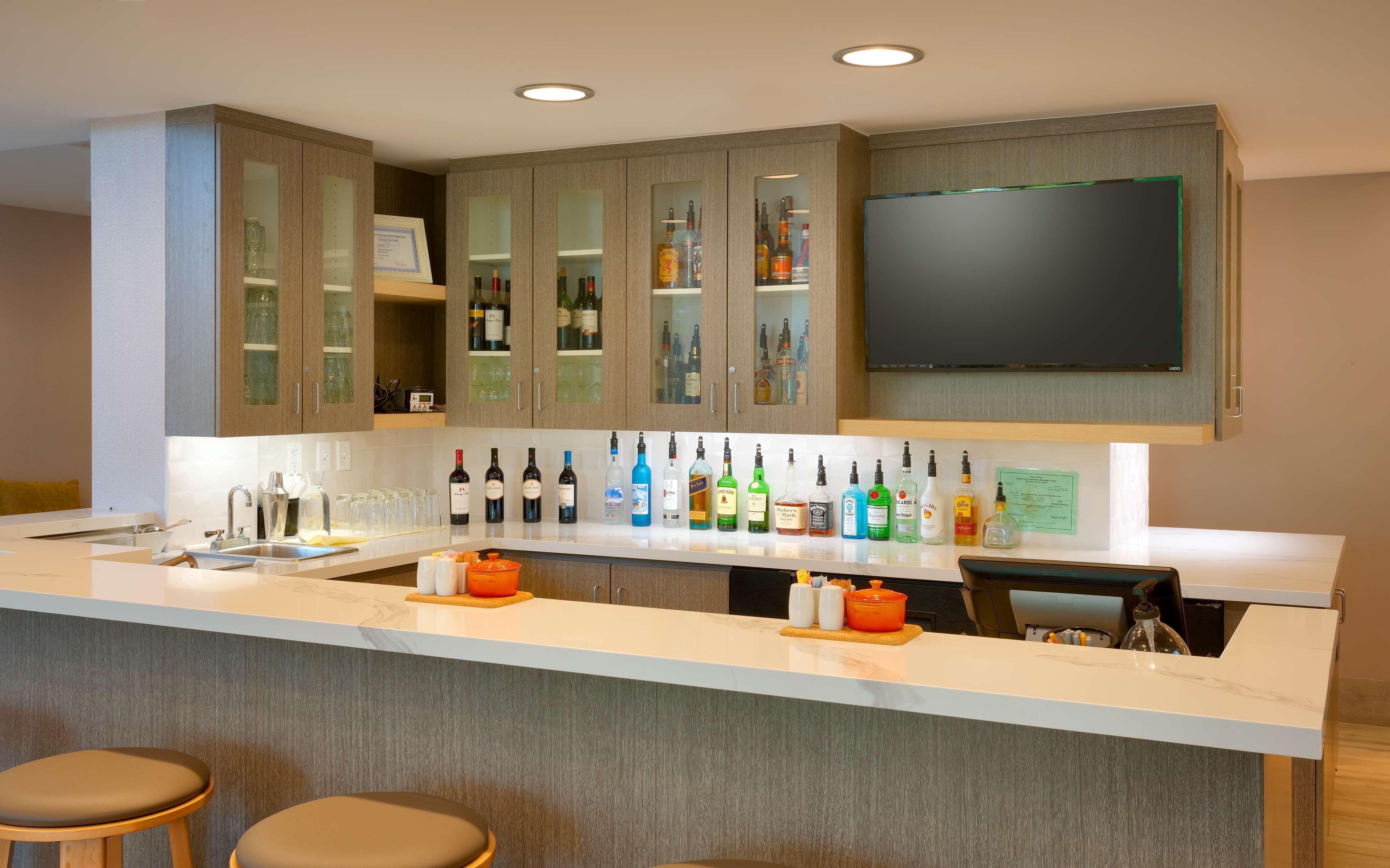 A modern kitchen with a bar area, featuring a wine and liquor display, a large flat-screen TV, and bar stools.