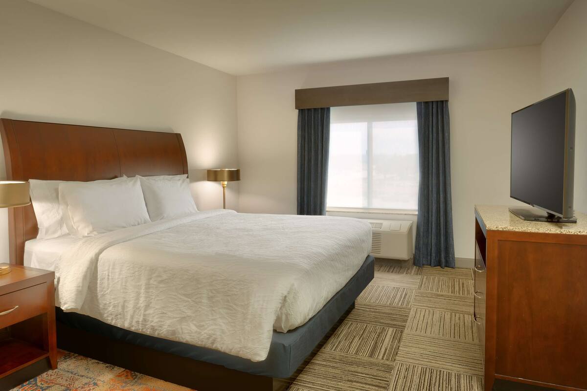 A hotel room with a large bed, a flat-screen TV, a desk with a lamp, and a window with curtains.