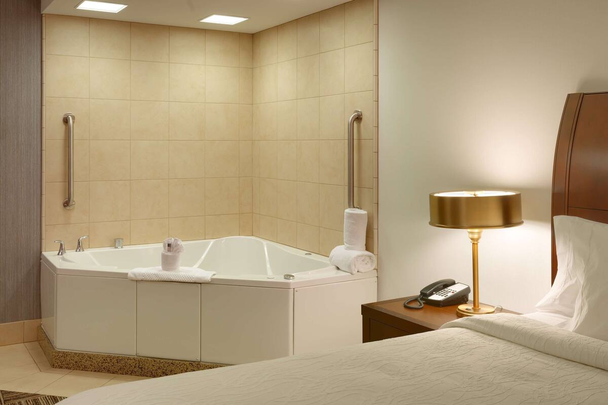 A hotel room with a corner bathtub, a bedside table with a lamp, and a bed with white linens.