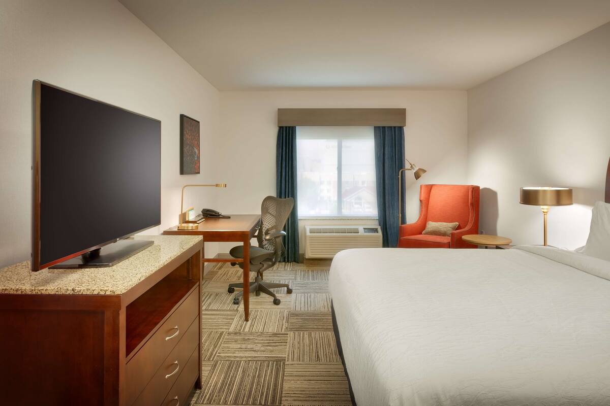 A hotel room with a large bed, a desk with a flat-screen TV, a chair, a small table, and a window with curtains.