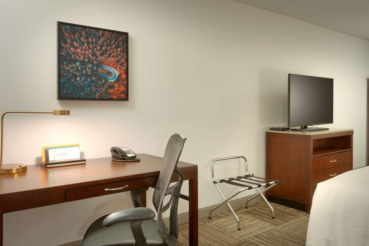 A hotel room with a desk, chair, TV, and artwork on the wall.