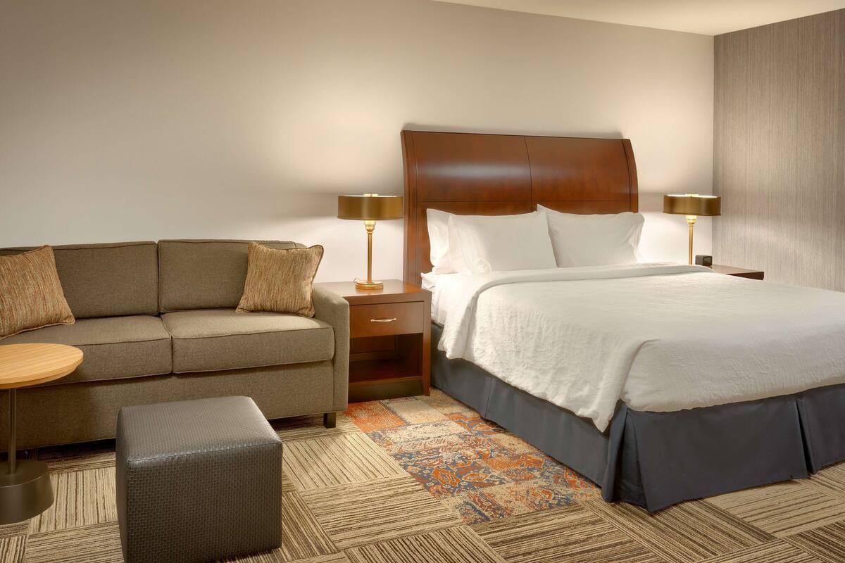 A hotel room with a bed, a sofa, a nightstand, and a lamp.