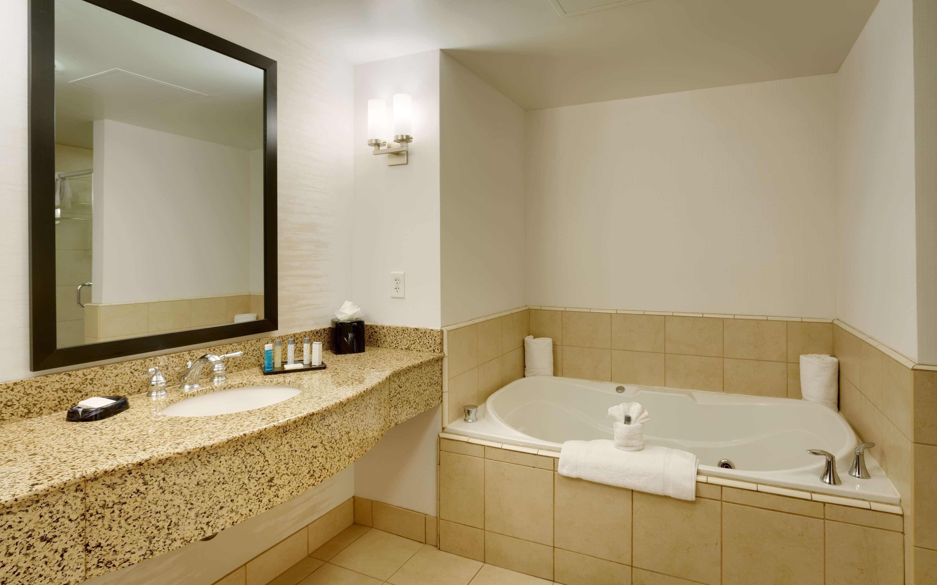 A bathroom with a granite countertop, two sinks, a large mirror, a wall-mounted light, a bathtub, and a towel rack.