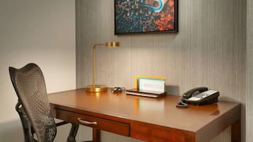 A hotel room with a wooden desk, a modern chair, a desk lamp, a telephone, and a framed artwork on the wall.