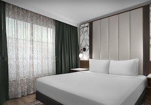 A modern hotel room with a large bed, a nightstand, a wall-mounted lamp, and a window with patterned curtains. - Embassy Suites by Hilton Jackson North Ridgeland (Ridgeland)