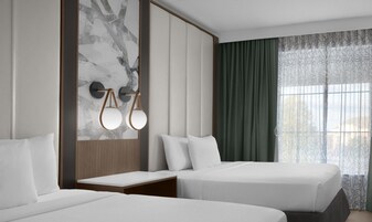 Habitación de hotel con dos camas, pared de mármol, cabecero de madera, mesita de noche y ventana con cortinas.