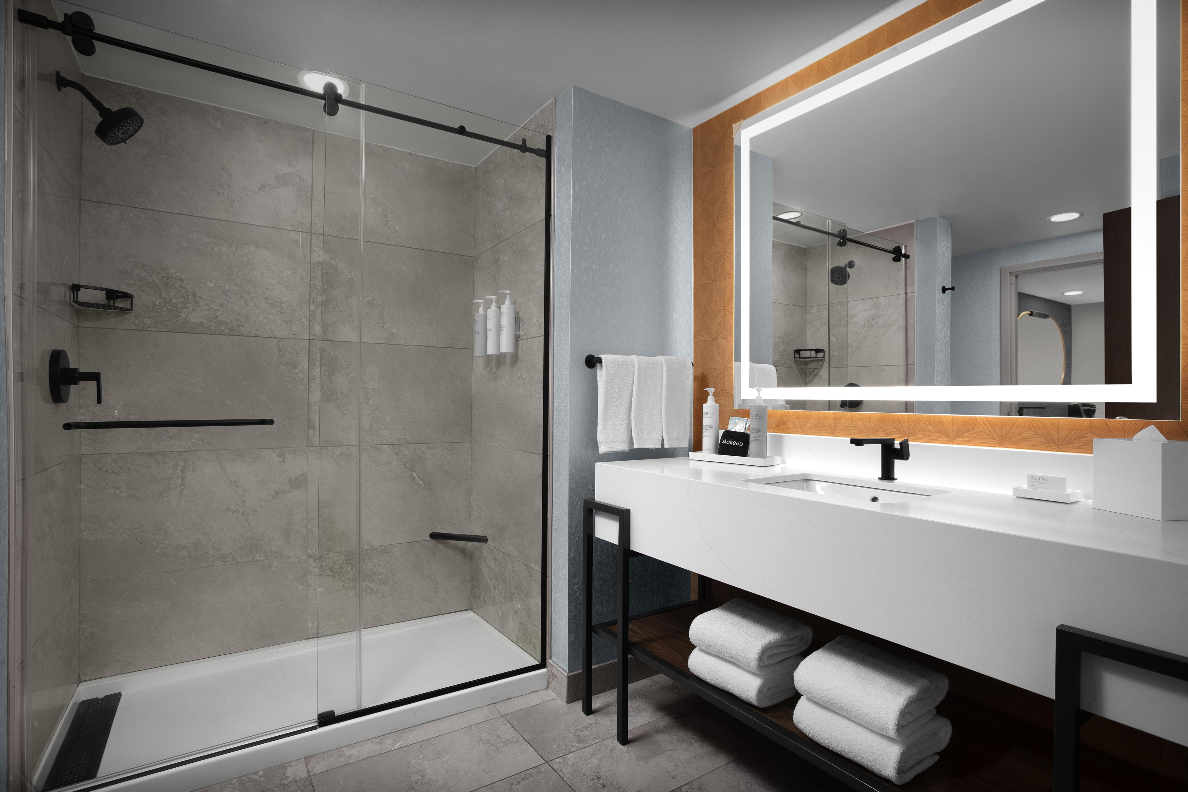 A modern bathroom with a glass-enclosed shower, a white vanity with a large mirror, and neatly folded towels on a shelf.