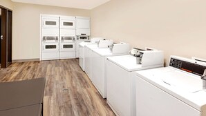 A laundry room with multiple white washers and dryers, wooden flooring, and a door on the left. - WoodSpring Suites Sioux Falls (Sioux Falls)