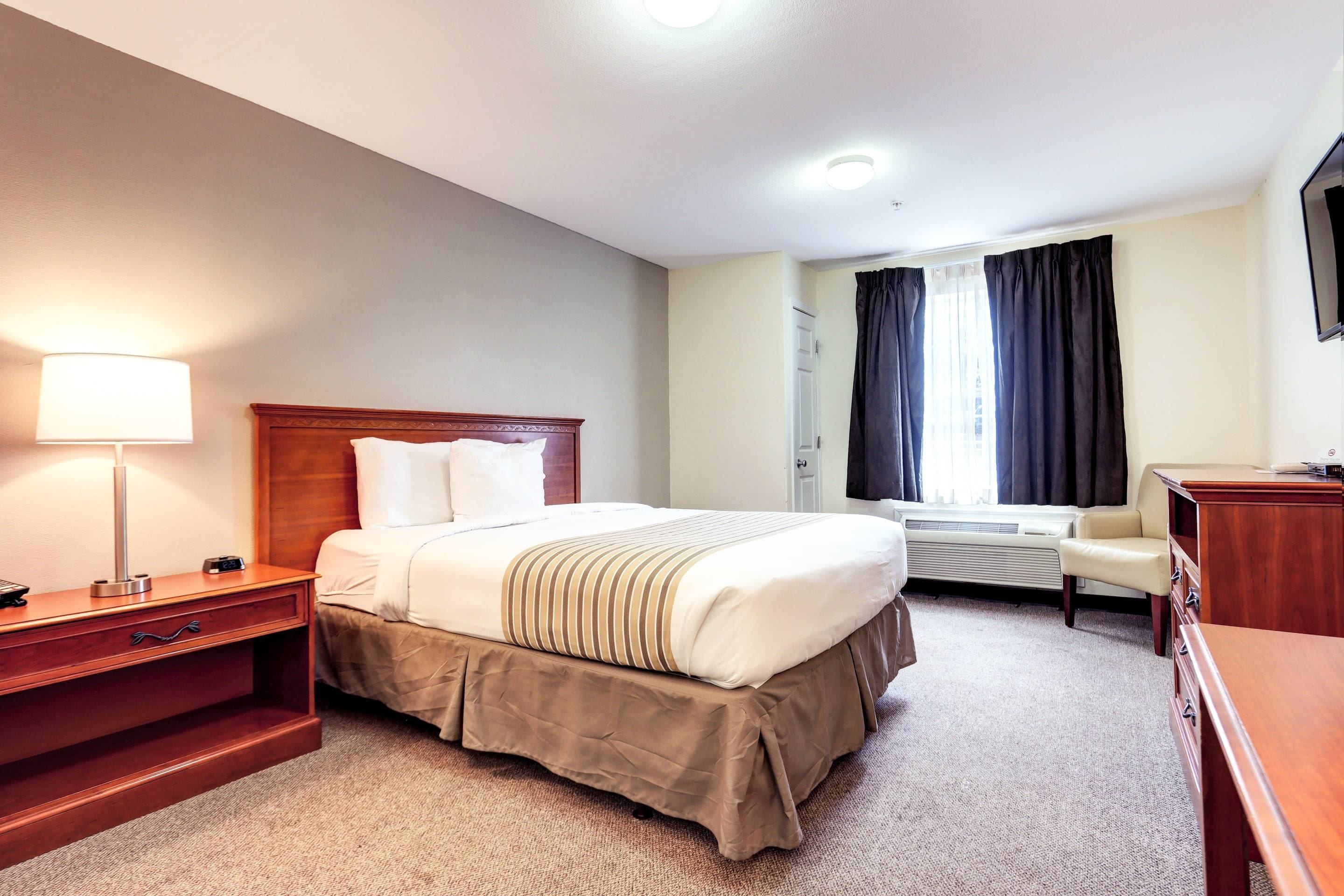 Une chambre d’hôtel comprenant un lit, une table de chevet, une lampe, une chaise, un bureau et une fenêtre avec des rideaux.