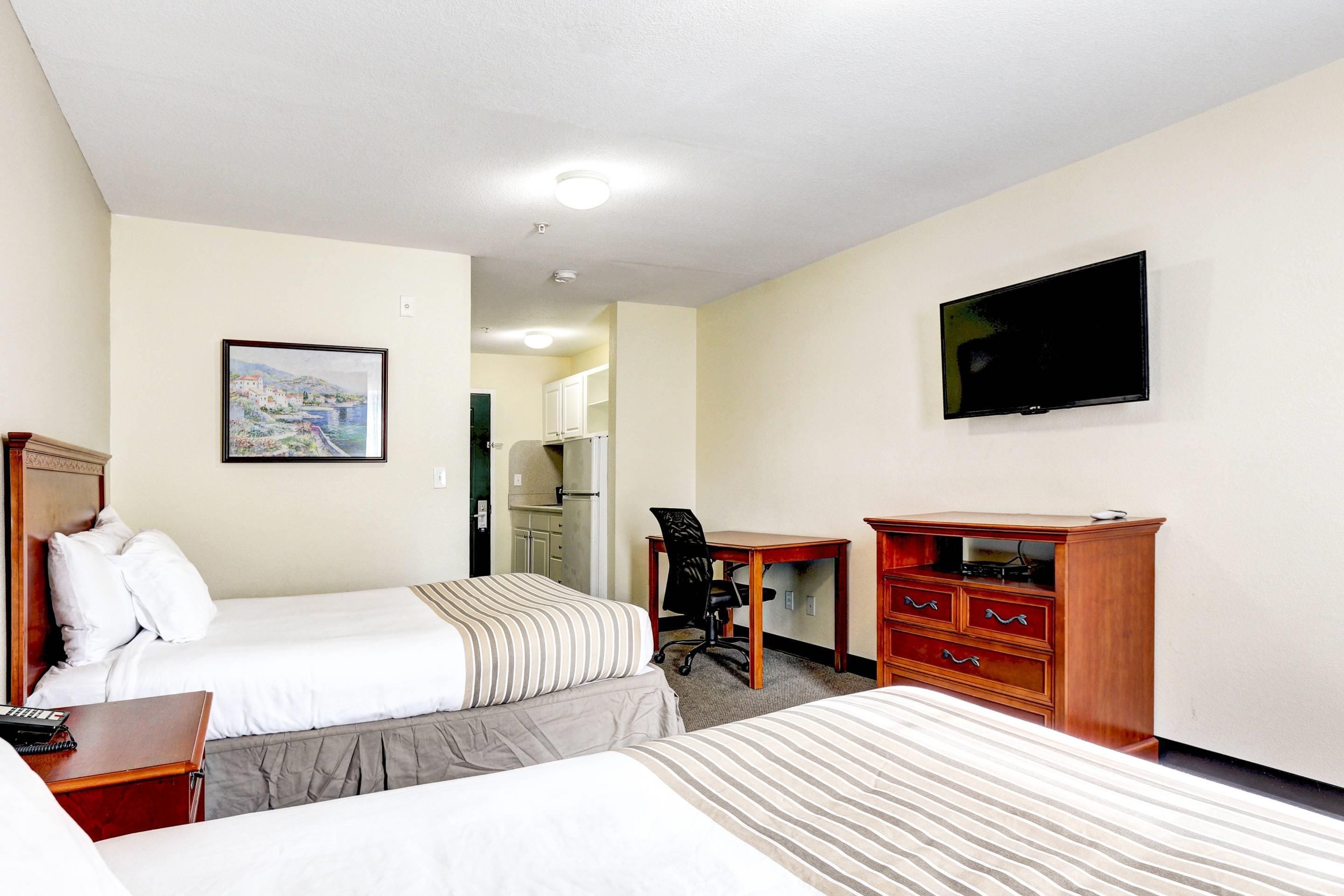 Quarto de hotel com duas camas, uma escrivaninha, uma cadeira, uma televisão e um quadro na parede.