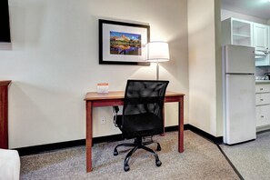 A hotel room with a desk, chair, lamp, and a refrigerator.