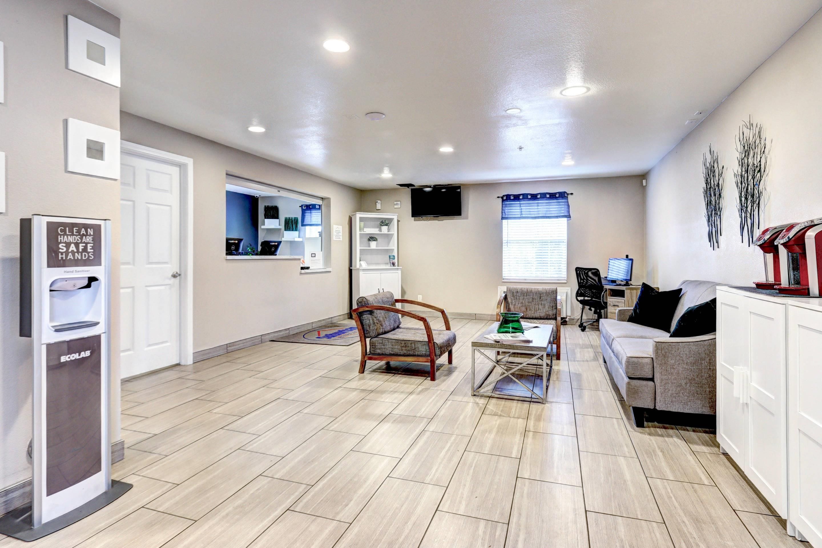 A clean, well-lit reception area with a hand sanitizer dispenser, a seating area with a sofa and chairs, and a television mounted on the wall.