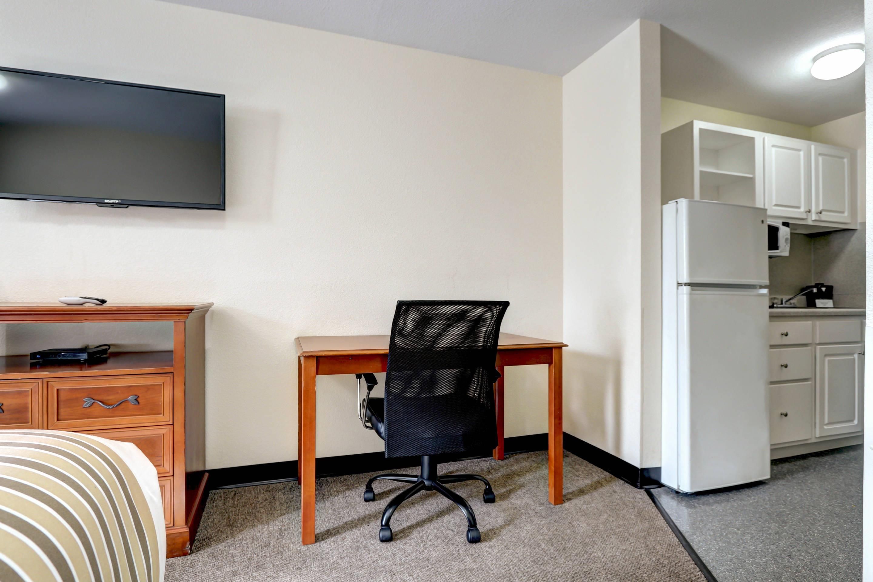 A hotel room with a wooden desk, a black mesh office chair, a flat-screen TV, a kitchenette with a white refrigerator, and a microwave.