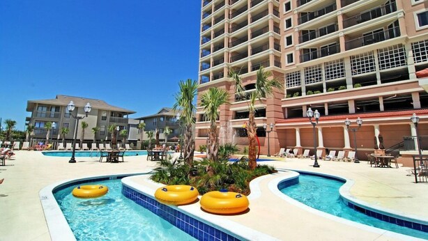 A hotel pool area with multiple swimming pools, lounge chairs, and palm trees. - Tilghman Beach And Golf Resort (North Myrtle Beach)