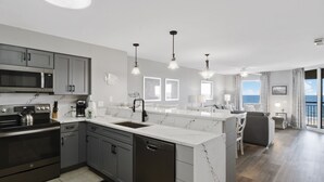 A modern kitchen with grey cabinets, a black stove, and a white marble countertop. There is a dining area with a white table and chairs, and a view of the ocean through large windows. - Tilghman Beach And Golf Resort (North Myrtle Beach)