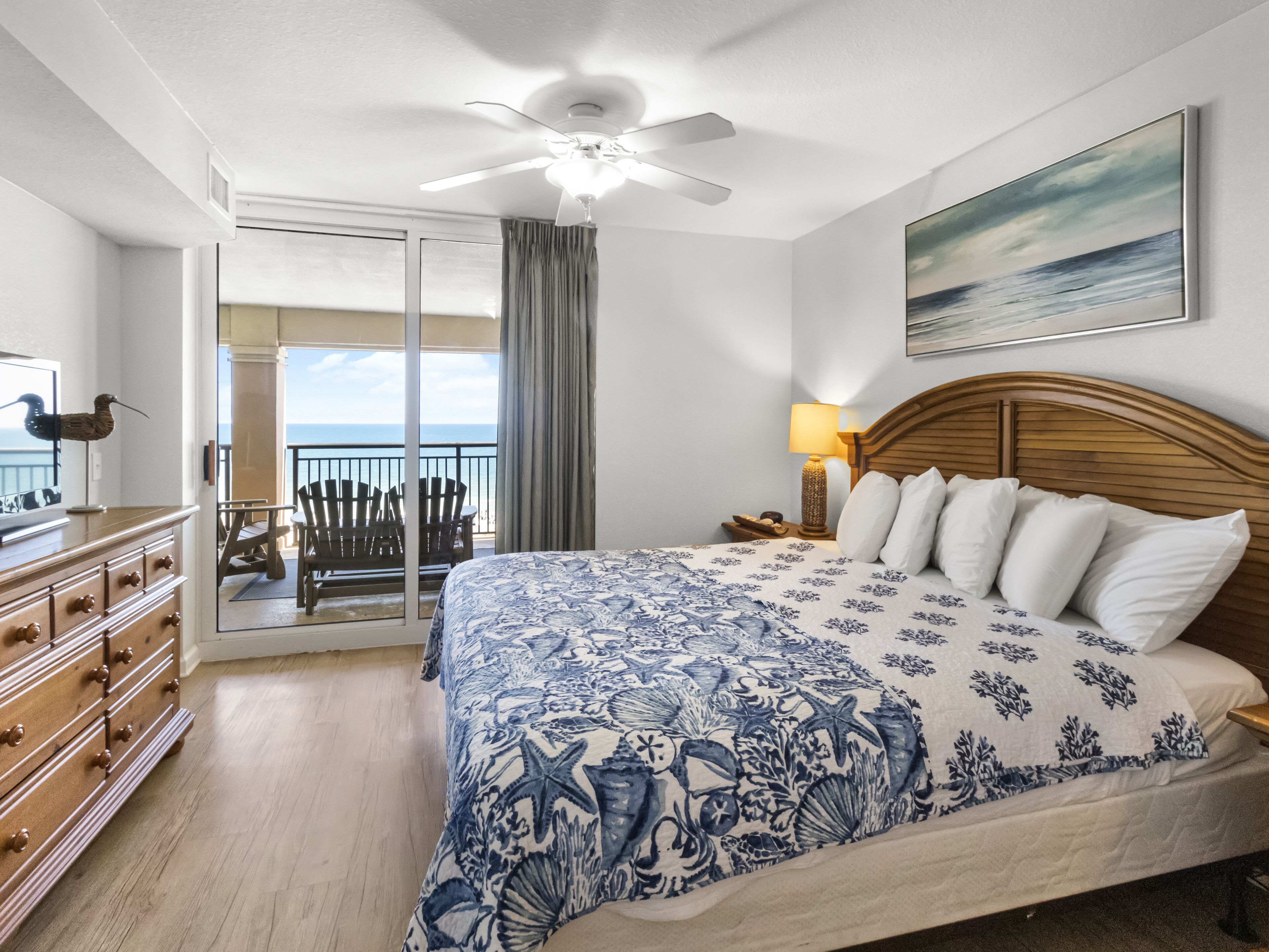 A bedroom with a large bed, a wooden dresser, a nightstand, a ceiling fan, and a view of the ocean through a sliding glass door.