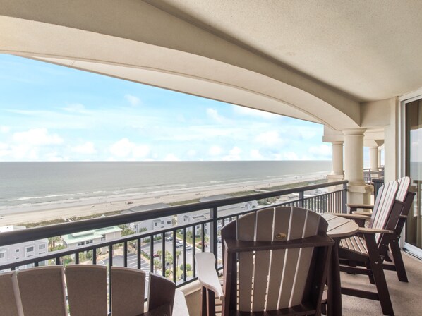 A balcony with a view of the beach and ocean. There are two Adirondack chairs and a small table.