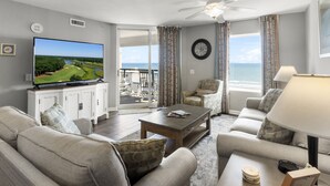 A living room with a television, a ceiling fan, a sofa, a coffee table, and a view of the ocean through large windows. - Tilghman Beach And Golf Resort (North Myrtle Beach)
