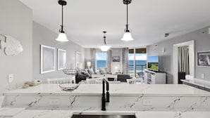 A modern kitchen with a central island, white marble countertops, and a stainless steel sink. There are pendant lights above the island and a large window offering a view of the sea. - Tilghman Beach And Golf Resort (North Myrtle Beach)