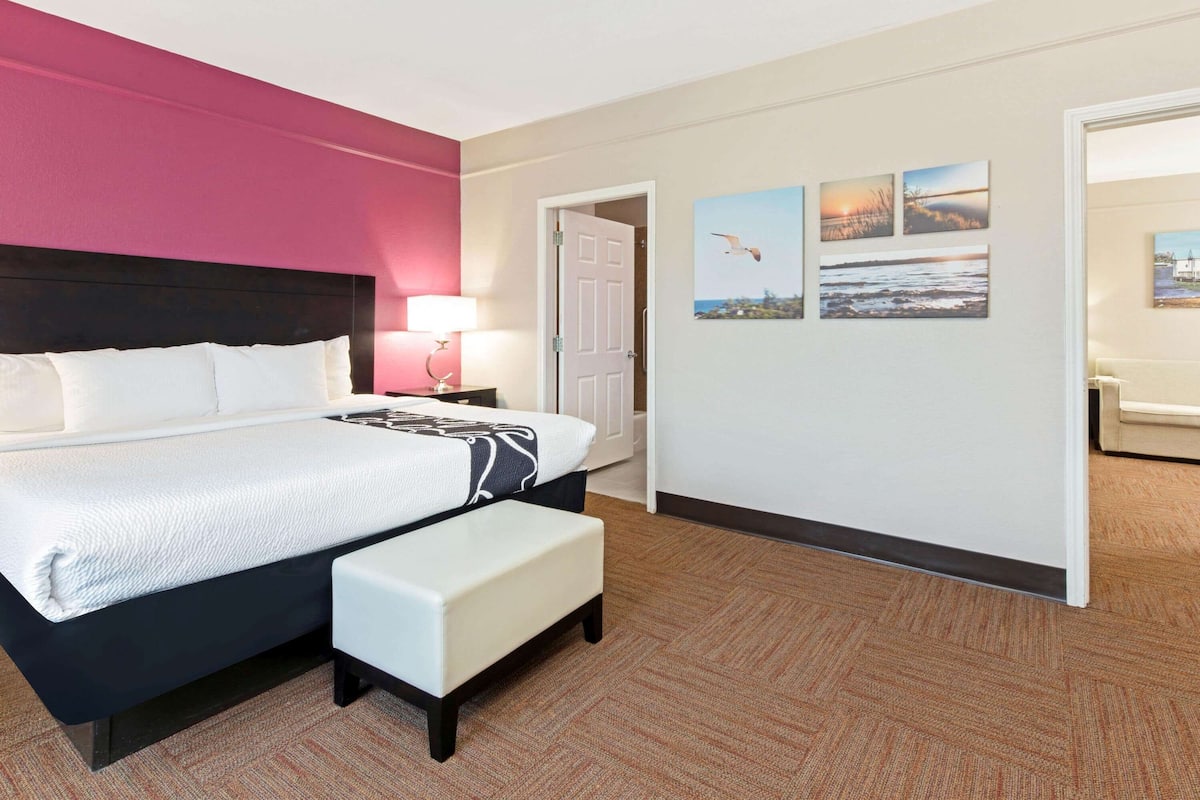 A hotel room with a large bed, a bedside table with a lamp, a bench, and a door leading to another room.