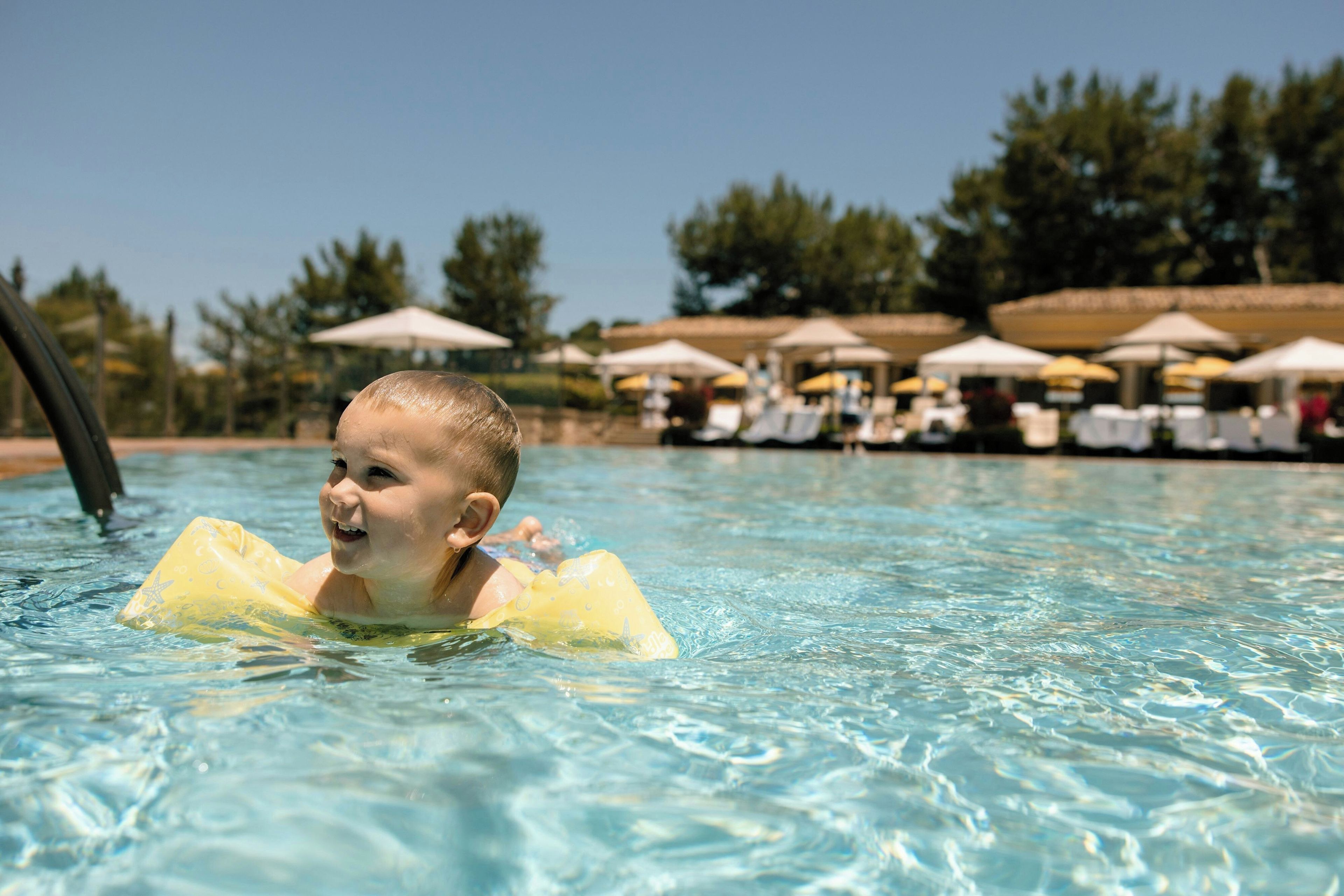 A child in a pool with clear water, surrounded by lounge chairs and umbrellas.