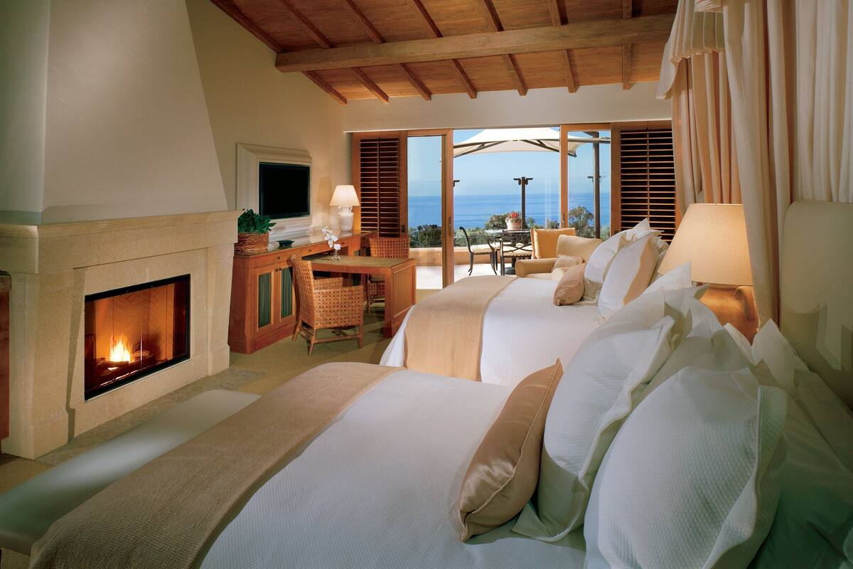 Ein Hotelzimmer mit zwei Betten, einem Kamin, einer Essecke mit Tisch und Stühlen sowie Meerblick durch große Fenster.
