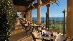 A restaurant with a long table, chairs, hanging baskets, and a view of the ocean. - The Resort at Pelican Hill (Newport Beach)