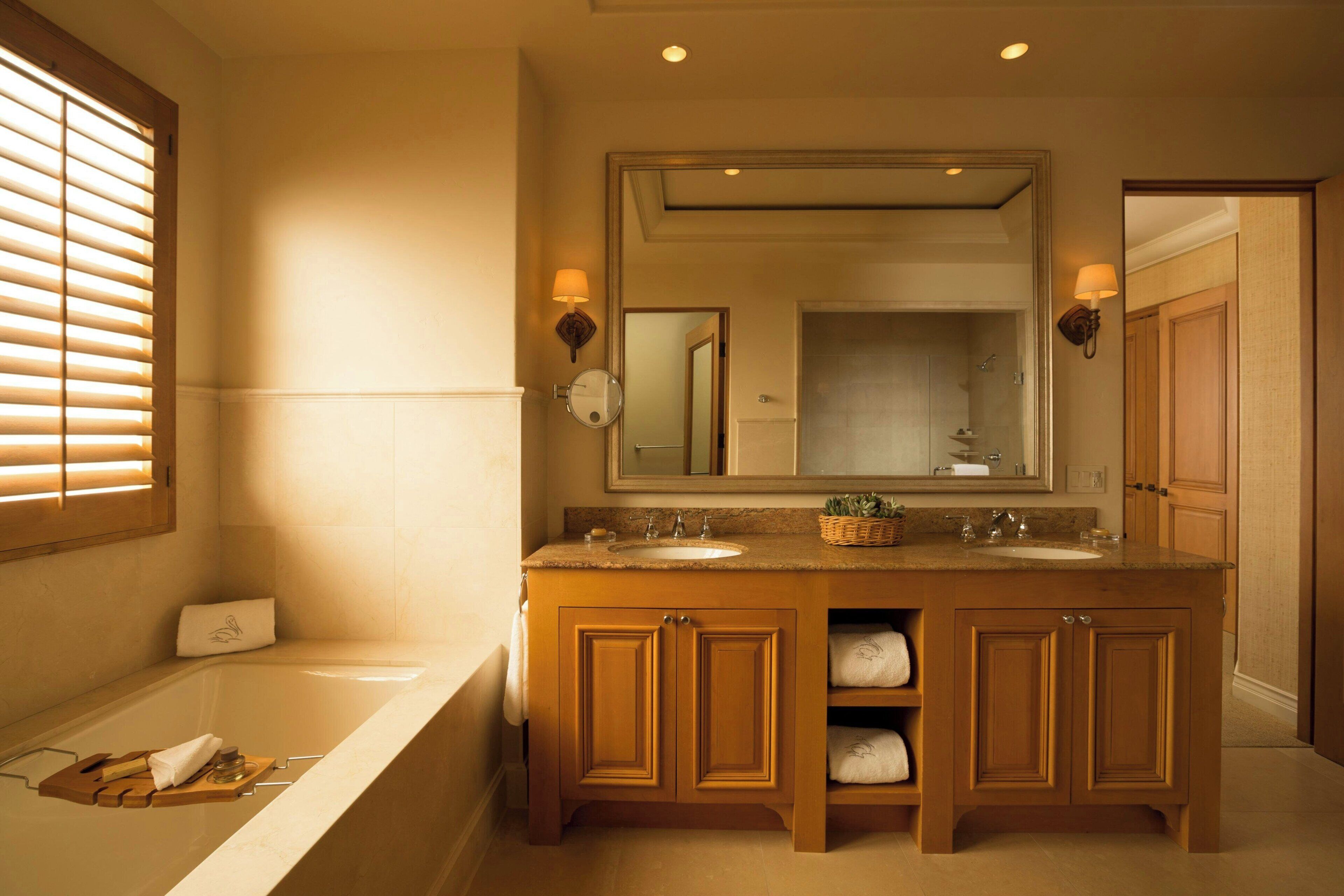 A bathroom with a bathtub, two sinks, wooden cabinets, a large mirror, and a window with blinds.