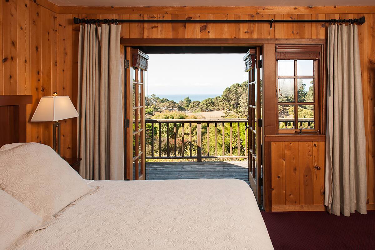 A wooden room with a bed, a lamp, and a view of a balcony with a wooden railing and greenery.